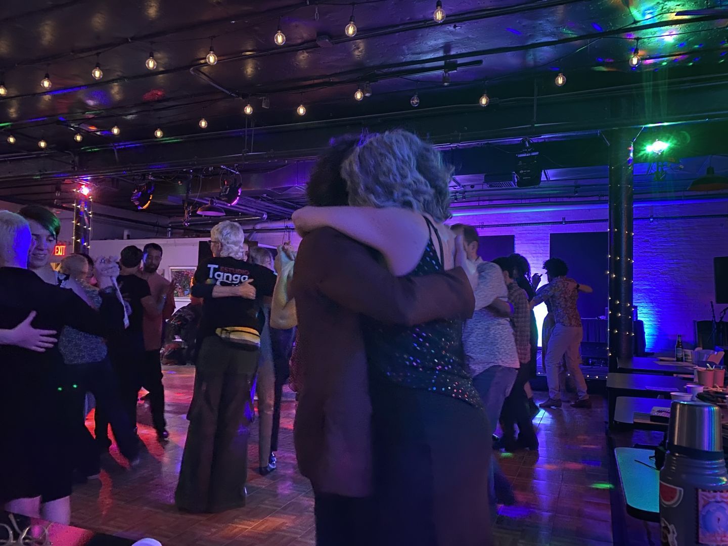 People in close embrace on a dance floor with string lights overhead. About 14 are visible, but there are more behind them. The closest to the camera is a person with shoulder length grey curls and a sparkly top held by someone in a maroon blazer.