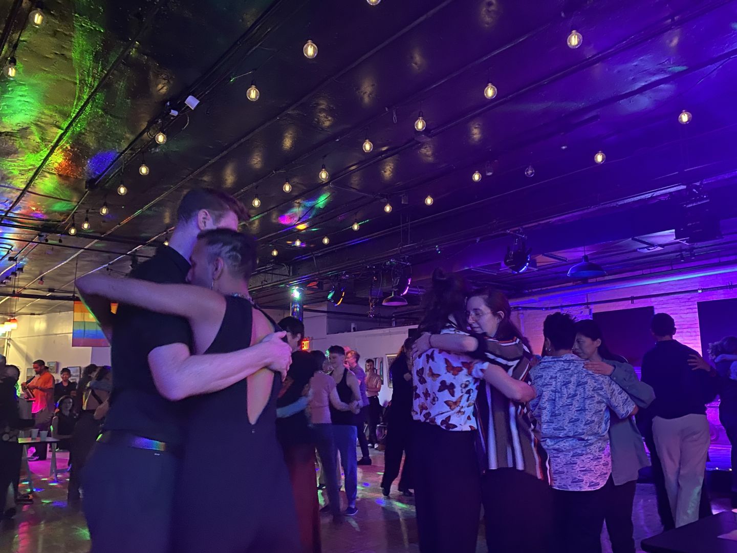 Couples embrace on the dance floor below string lights with a pride flag in the background