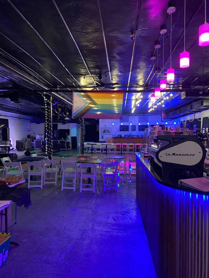 An open space with a dance floor surrounded by chairs. A progress Pride flag hangs from the ceiling. An espresso bar is in the foreground. A stage with a baby grand piano is on the left.