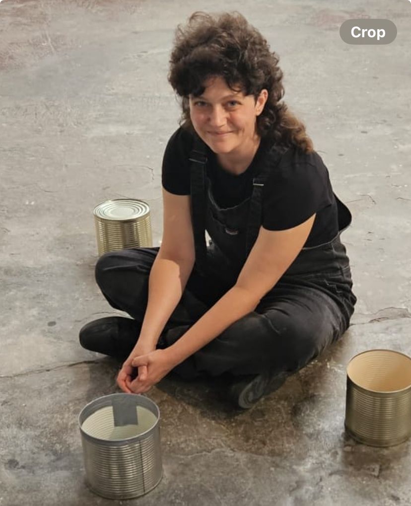 white non binary person, dressed in black dungarees and black t shirt, sitting cross-legged on concrete floor looking up at camera smiling, surrounded by 3 large tin cans on floor.